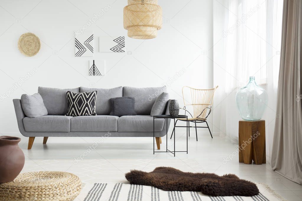Real photo of a brown, fur rug lying on white floor in front of a gray couch with cushions in bright living room interior