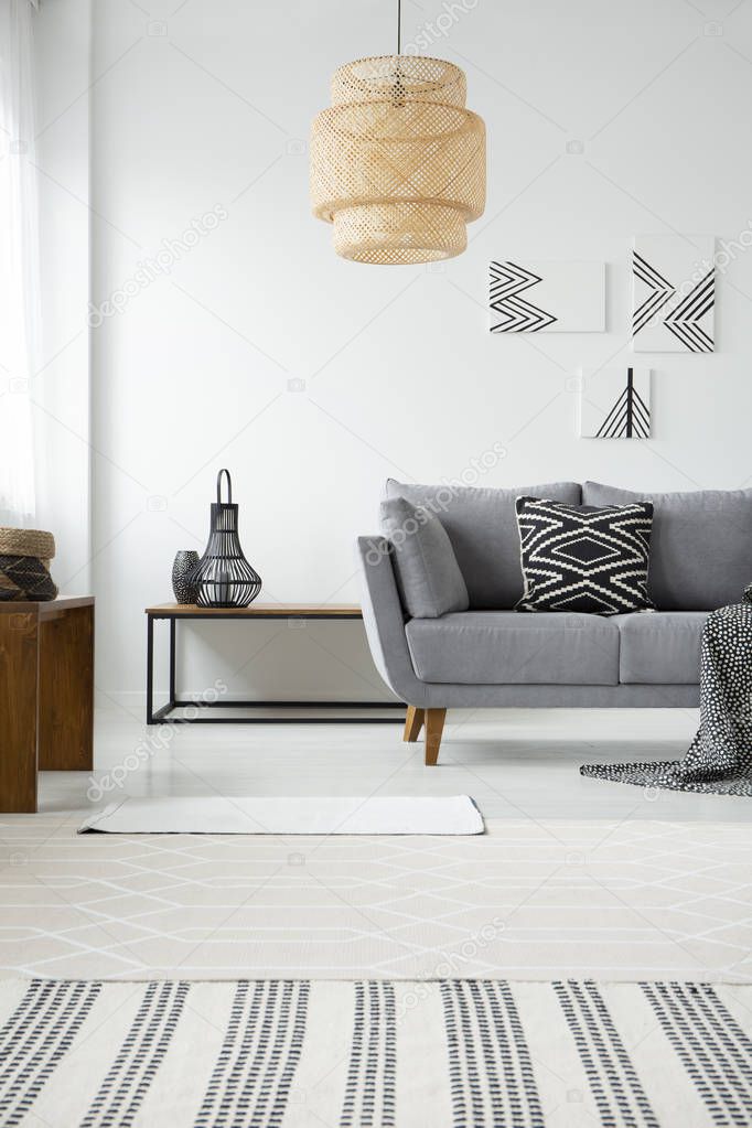 Real photo of a rattan lamp hanging above a gray settee with a black and white cushion in scandi living room interior