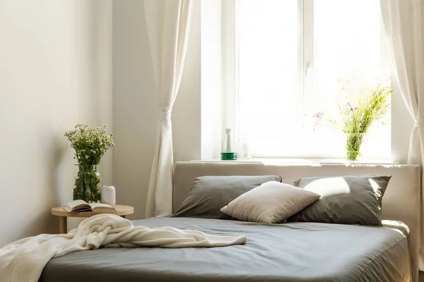 毯子和枕头在灰色床在明亮的卧室内部与花和窗口 真实照片 — 图库照片