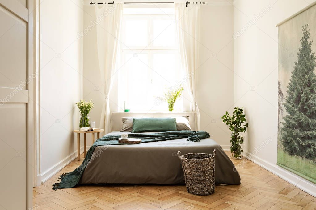 Green blanket on bed and plants in bedroom interior with window and tree painting. Real photo
