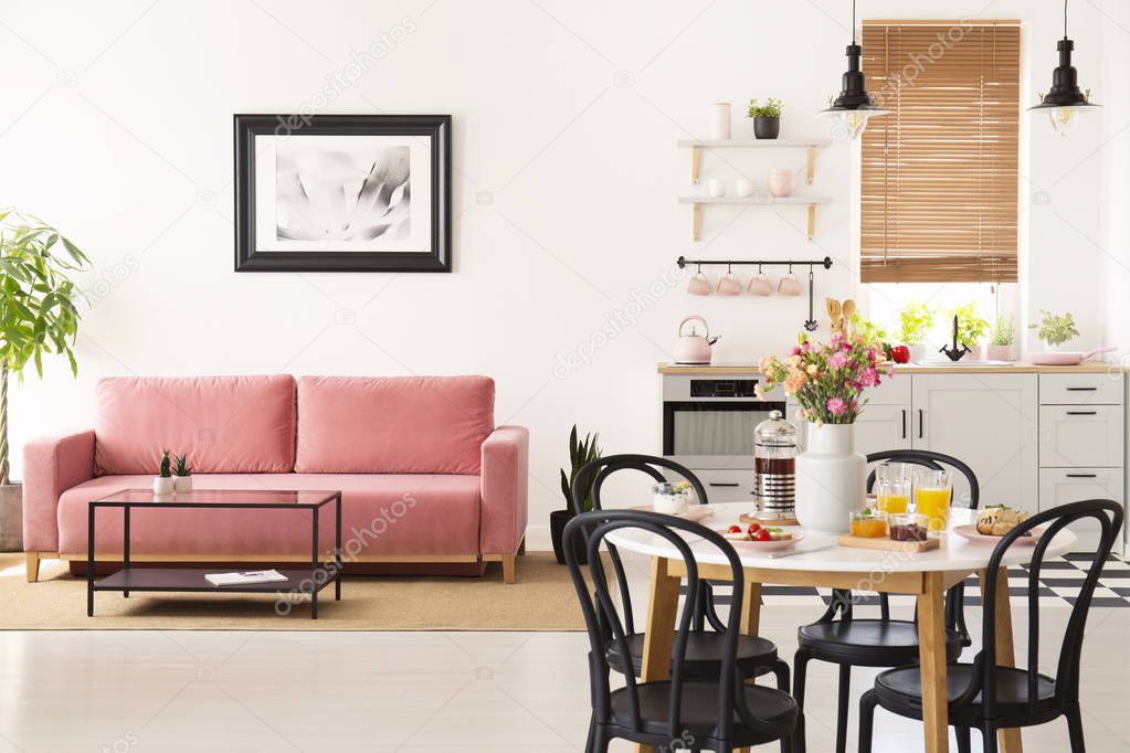 Dining table with breakfast and fresh flowers standing in bright apartment interior with pink sofa, black and white poster and kitchenette with pastel accessories