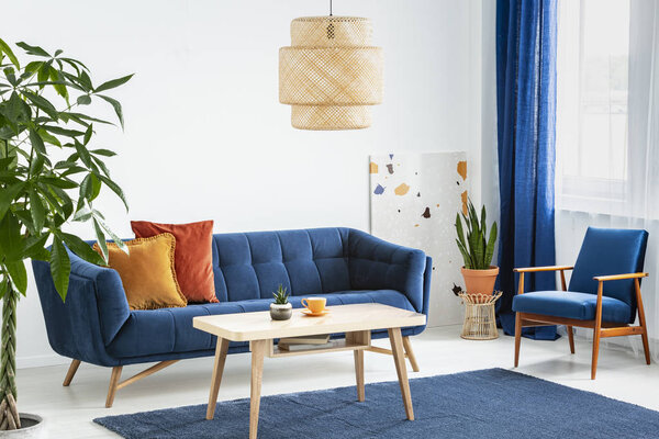 Armchair and sofa in blue and orange living room interior with lamp above wooden table. Real photo