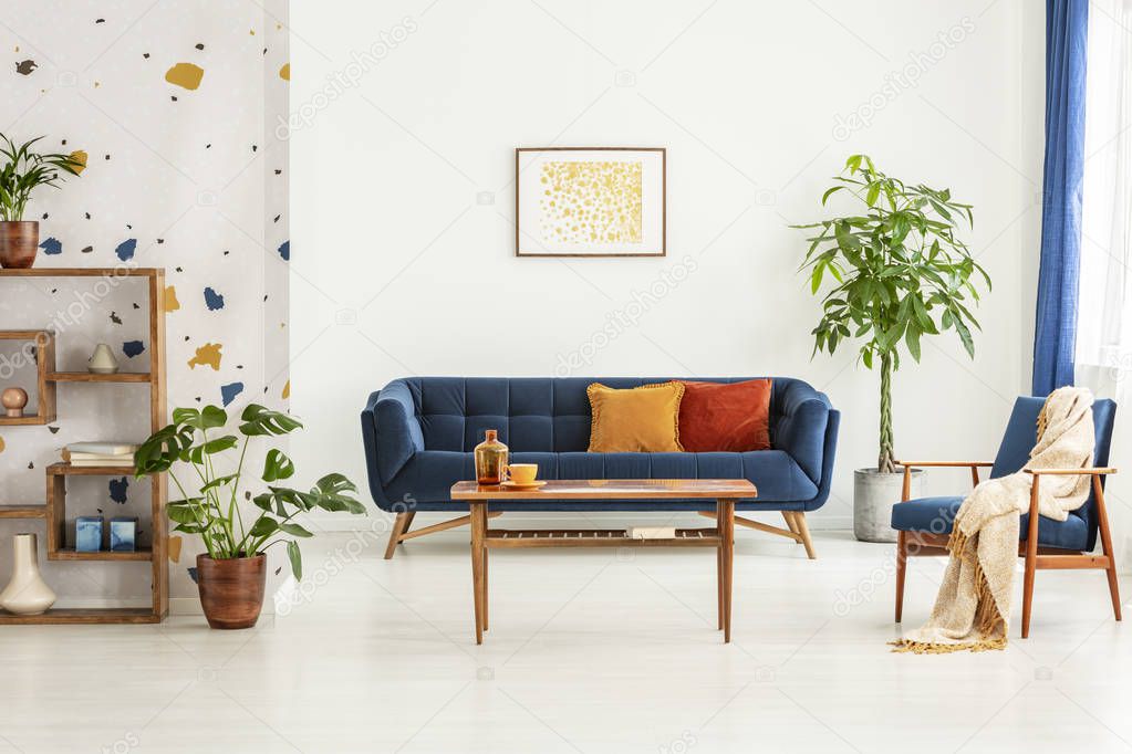 Poster above blue settee in white apartment interior with armchair, wooden table and plants. Real photo