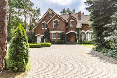 Large cobbled driveway in front of an impressive red brick English design mansion surrounded by old trees clipart