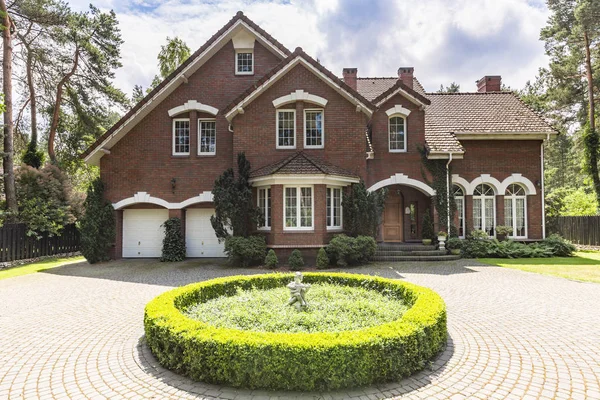 Front view of a red brick English style classic house with a steep roof, large windows and a circular driveway with a flowerbed decoration in the front.