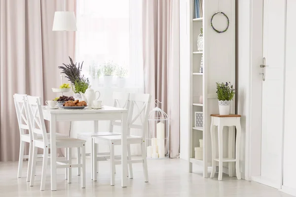 White chairs at dining table with plants in modern flat interior with pink drapes and window. Real photo
