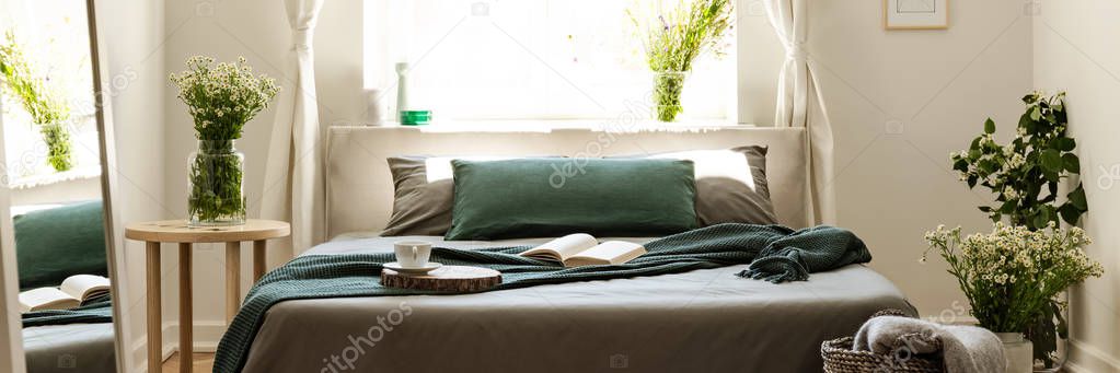 Bright bedroom interior in real photo with fresh flowers, window and double bed with open book, tea cup and green blanket