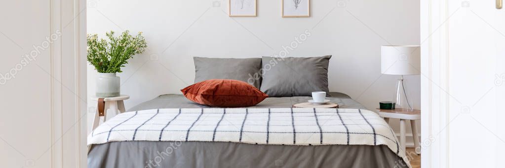 Real photo of king-size bed with grey sheets, coffee cup, striped blanket and velvet cushion standing in white bedroom interior with fresh flowers and glass lamp