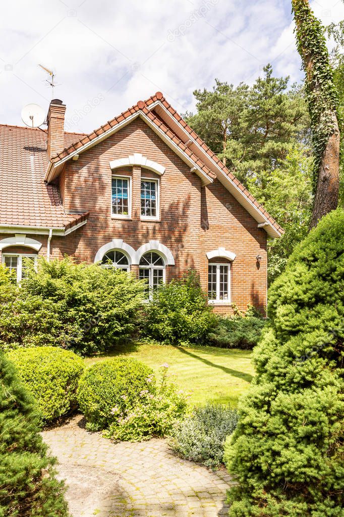 A garden with evergreens, trees, shrubs and grass in the backyard of a red brick luxurious English style house.
