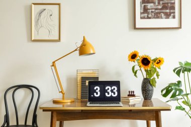 Sunflowers, yellow lamp and laptop on wooden desk in home office interior with posters. Real photo clipart