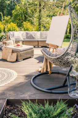 Hanging chair on wooden patio with easel and rattan table in front of sofa in the garden clipart