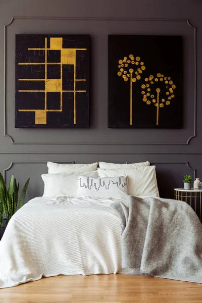 Black and gold posters above white bed with blanket in grey bedroom interior. Real photo