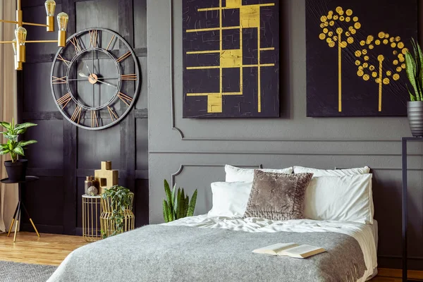 Black and gold posters above bed in grey bedroom interior with clock and plants. Real photo