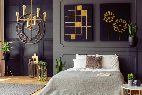 Plants and black and gold posters in grey bedroom interior with clock and bed with pillows. Real photo