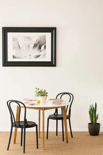黒い椅子のジュース パステル ピンク プレート 新鮮な植物の壁にポスターと白い部屋インテリアの本物の写真でケーキと木製のダイニング テーブルのそばに立って — ストック写真