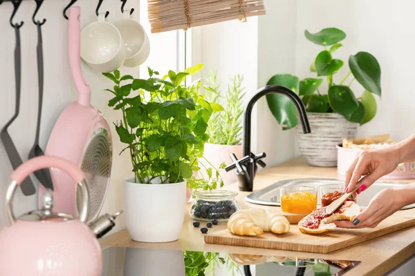 女性ピンク ポット 植物のキッチン インテリアの木製の机でパンにジャムを広がっています 実際の写真 — ストック写真