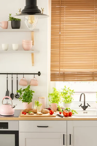 Real photo of bright kitchen interior with fresh plants, tomatoes and bread placed on countertop, window with wooden blinds and pastel pink utensils