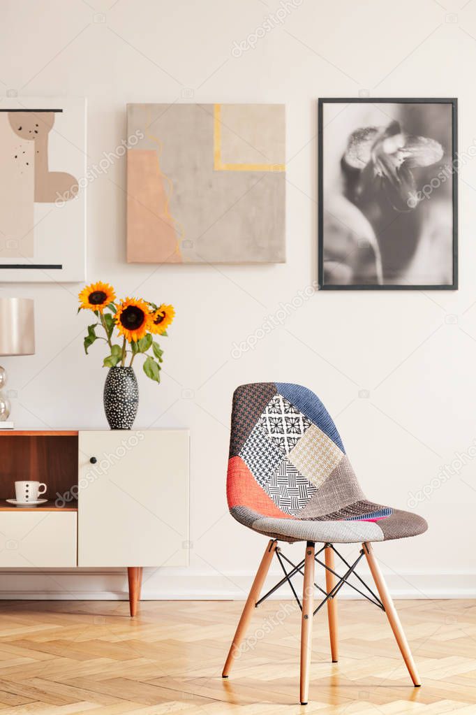 Modern chair in white apartment interior with posters above cupboard with sunflowers. Real photo