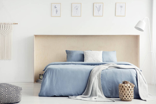 Blanket and pillows on blue bed in white bedroom interior with pouf, lamp and posters. Real photo