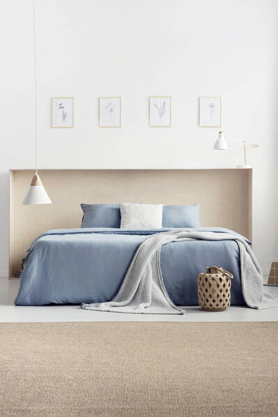 Одеяло и подушки на голубой кровати в светлом интерьере спальни с ковровым покрытием и плакатами. Настоящее фото
