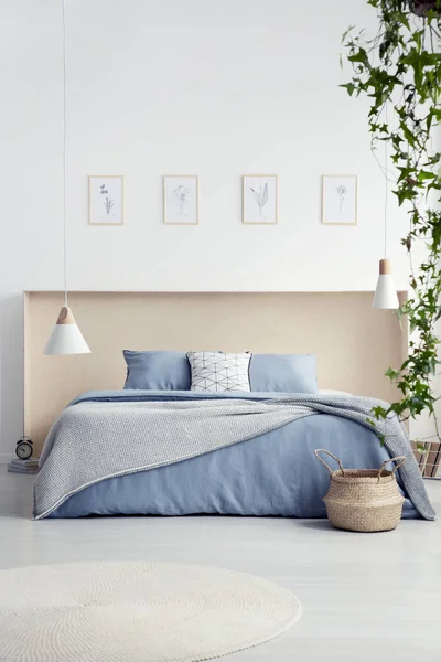 Round rug and lamp in white bedroom interior with posters above blue bed with blanket. Real photo