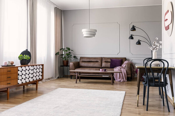 Wooden cupboard in bright living room interior with leather sofa and chairs at table. Real photo