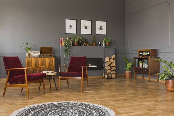 Round rug in front of red armchairs in grey retro living room interior with posters and radio. Real photo