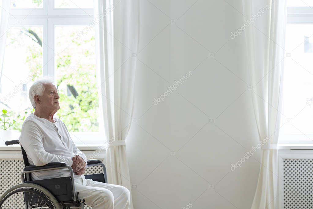 Lonely elderly patient in a wheelchair in a white room next to an empty wall. Place your logo