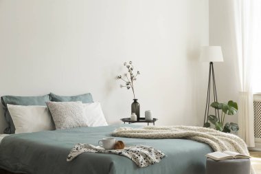 Bedroom interior with sage green and white sheets and cushions and a blanket. Black metal table with vases beside the bed. A lamp standing in the corner. Real photo.