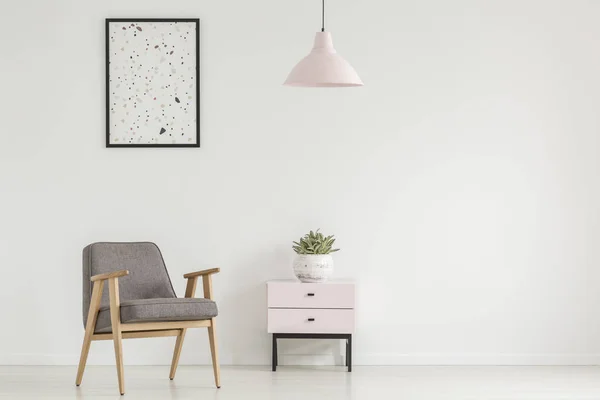 Poster above grey wooden armchair next to cabinet with plant in white flat interior with lamp. Real photo