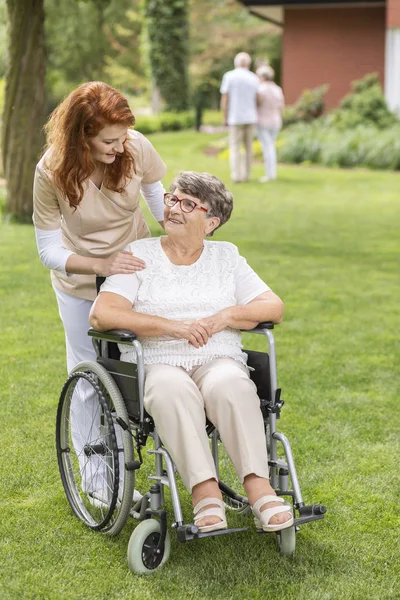 支援し 民間のリハビリ クリニックの庭に夏の日に車椅子で年配の女性患者の後ろに立って専門的な世話人 — ストック写真