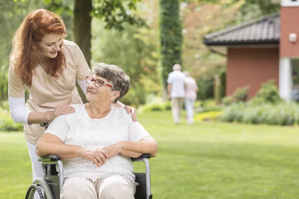 一名女性志愿者 在私人豪华养老院外的花园里 帮助残疾的老年妇女坐轮椅 背景模糊 — 图库照片