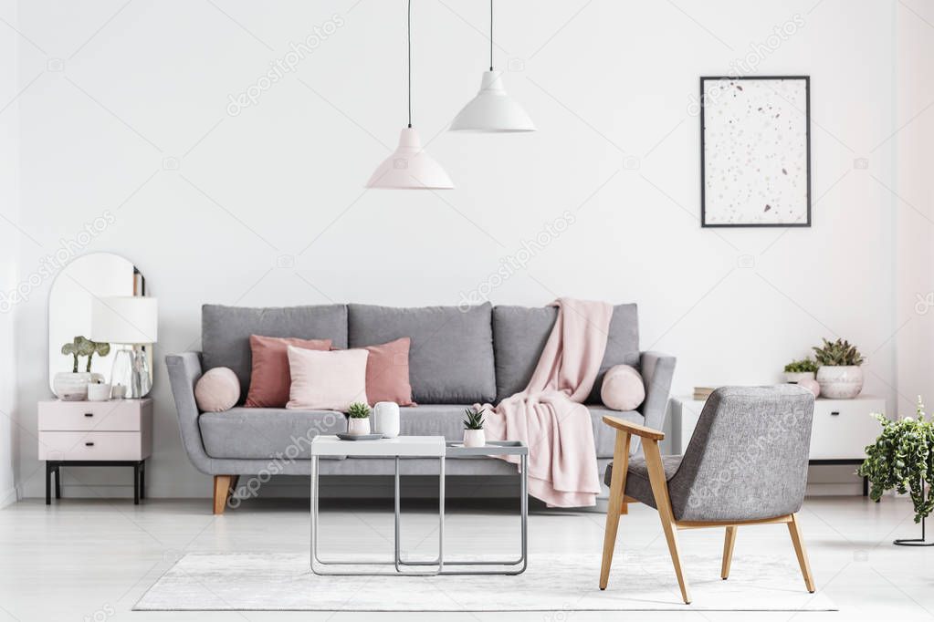 Grey armchair next to tables in white living room interior with poster and pillows on sofa. Real photo