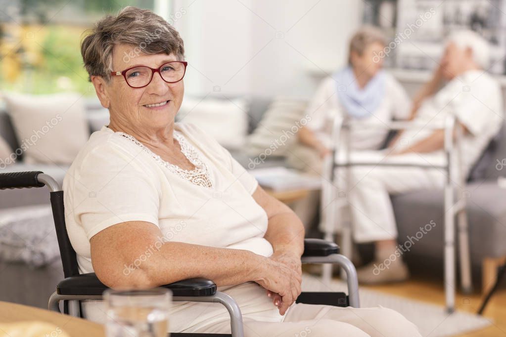 A portrait of a smiling elderly woman in a wheelchair inside a common room of a luxurious care home for seniors. Blurred background.