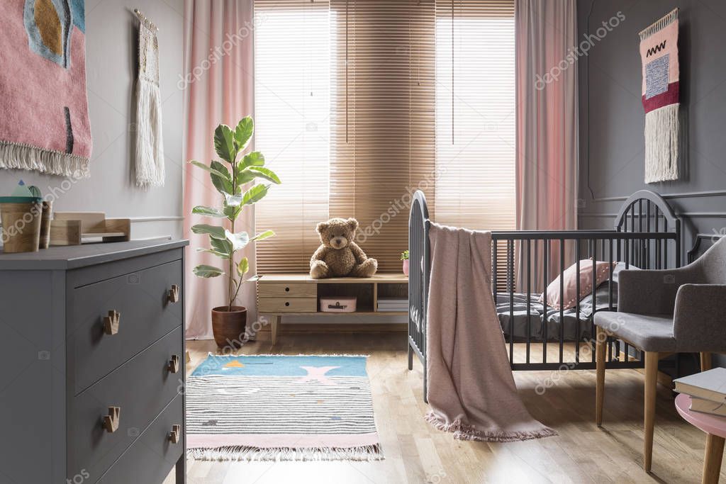 Teddy bear on wooden cupboard next to ficus in child's bedroom interior with cradle. Real photo