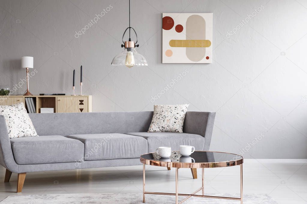 Lamp above grey settee with cushions in bright living room interior with poster and table. Real photo