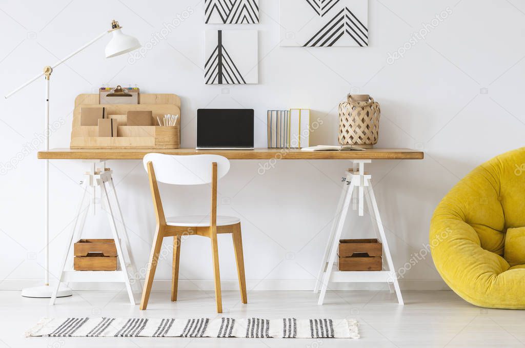 Yellow pouf next to desk and wooden chair in white home office interior with posters. Real photo