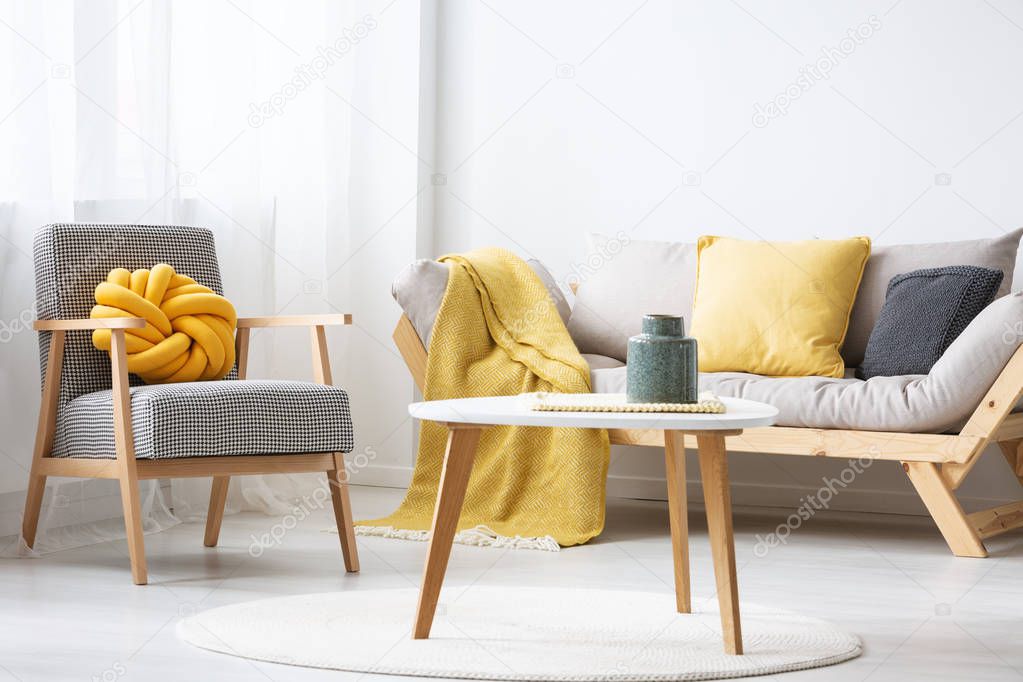 Patterned armchair next to settee with yellow blanket in flat interior with wooden table. Real photo