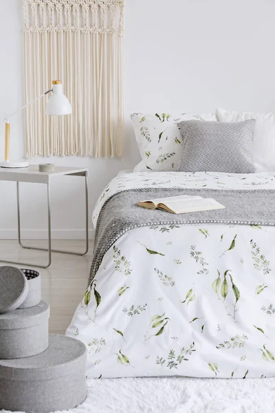 A cozy pastel bedroom interior with a bed dressed in green plants on white linen and cushions. Warm gray wool blanket on the bed. Real photo.