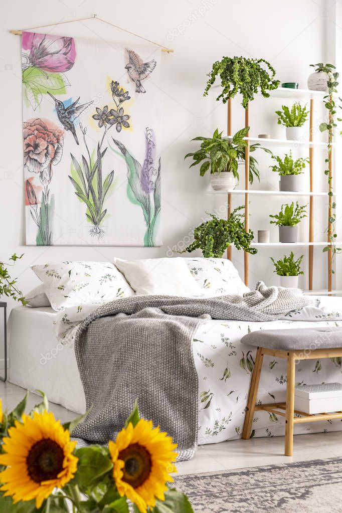 Urban jungle bedroom interior with sunflowers in the foreground and many green plants beside a bed dressed in white bedding. Tapestry on the wall. Real photo.