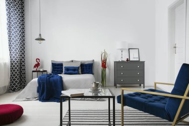 Bir yatak, Komidin, çiçek, sehpa, koltuk ve mavi aksan ile modern yatak odası iç gerçek fotoğraf. Duvar boş, tablonu yerleştirin