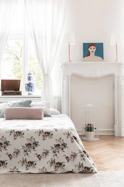 Bed with rose decorated cover and an ornamented fireplace mantel in a high ceiling vintage bedroom interior. Real photo.