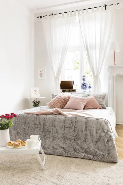 High ceiling bedroom interior with a bed dressed in a silver bedding and lemonade pink pillows and a blanket. Breakfast and roses on a tray standing on a fluffy rug. Real photo.
