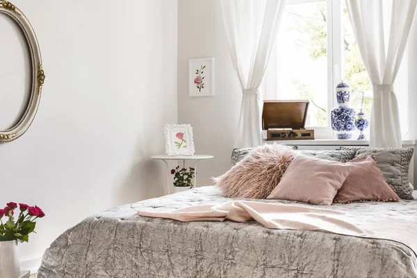 Soft gray and lemonade pink bedding and pillows on a bed which is standing against a bright window in an English style bedroom interior. Real photo.