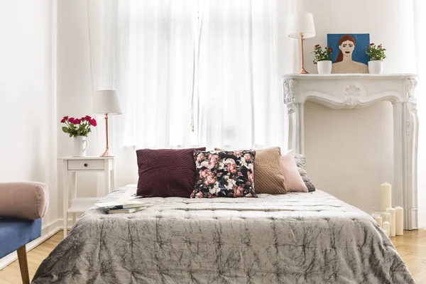 Chic bedroom interior featuring a bed with multicolor cushions and a velvet throw and white ornamented fireplace mantel as a decoration. Real photo.