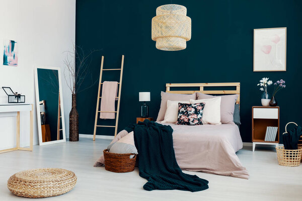 Dark blanket thrown on double bed with many pillows in real photo of bedroom interior with mirror, flowers on bedside table and poster on dark blue wall