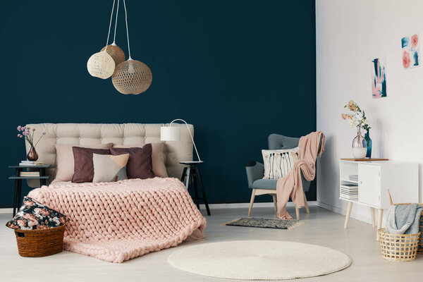 Grey armchair with powder pink blanket and cushion standing in the corner of white and blue bedroom interior with king-size bed with handmade coverlet and many pillows