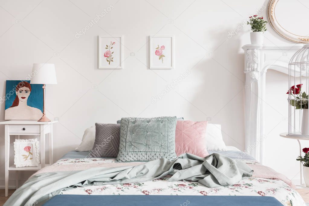 Pastel bedroom interior with roses as a pattern on bed linen and illustrations on the wall. Self portrait of a woman on a nightstand. Real photo.