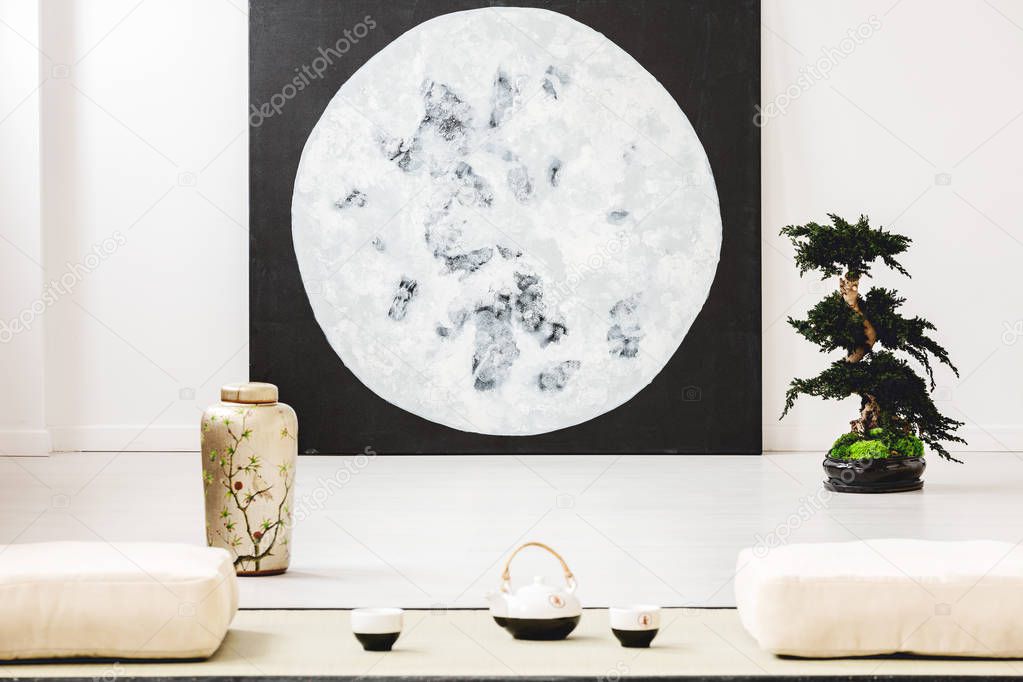 Moon poster next to bonsai in white interior in japanese style with pillows on the floor. Real photo