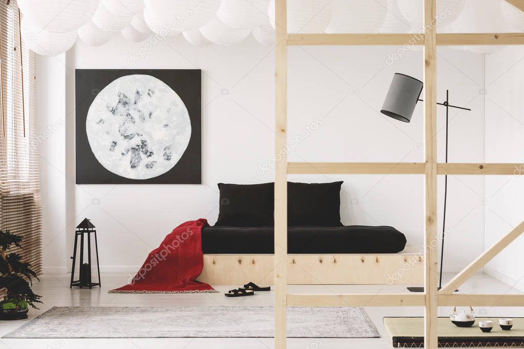 Red blanket and black cushions on wooden bed in bedroom interior with moon poster and tategu. Real photo
